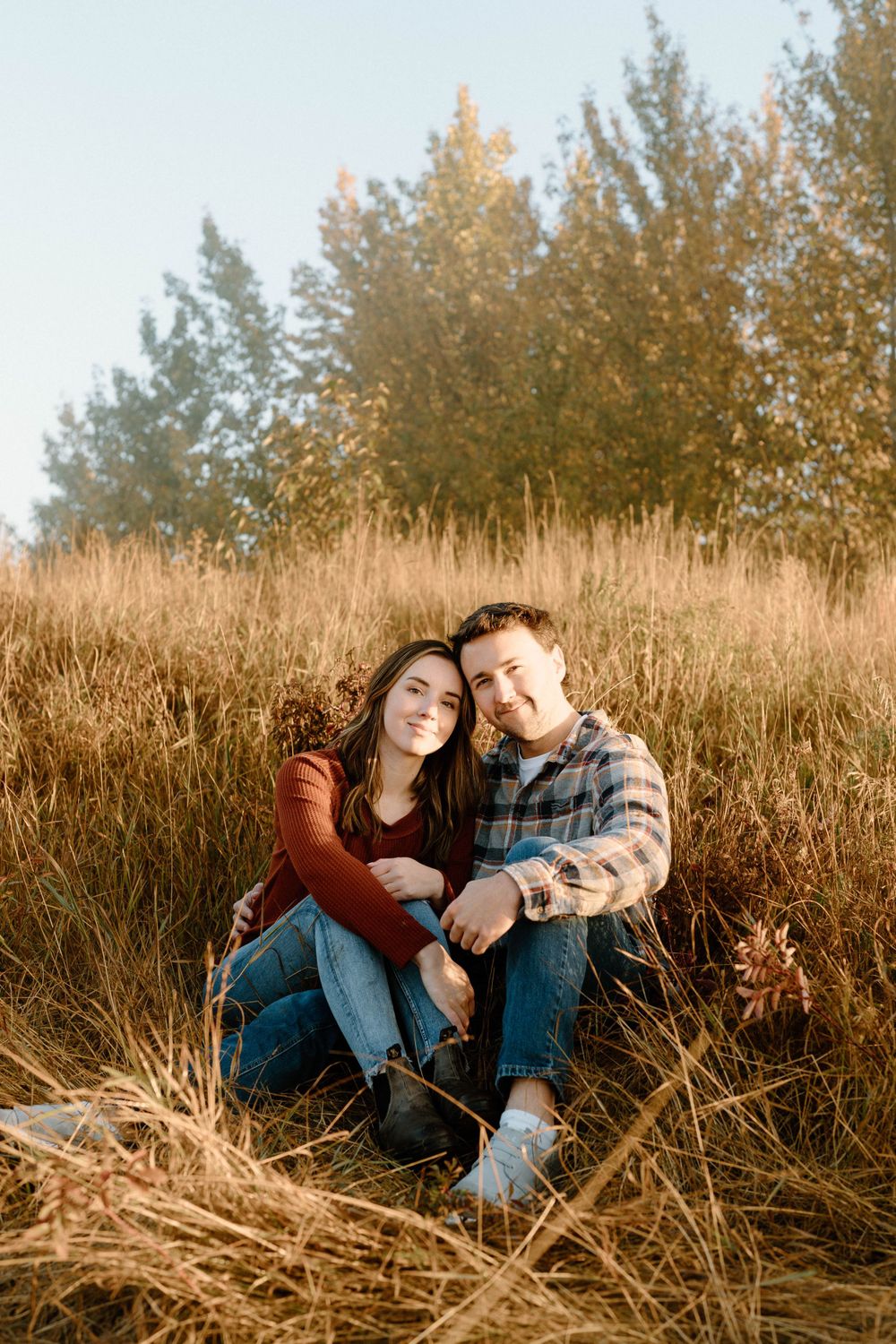 A couple in casual attire sits close together in autumn grass with trees behind them.