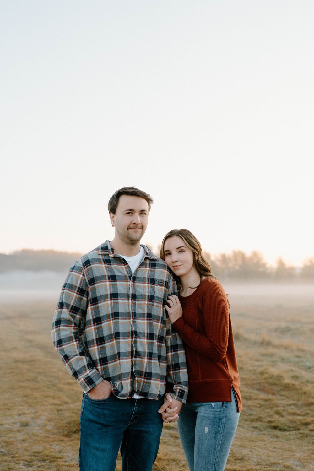 A couple in casual wear embraces in a misty field during golden hour.