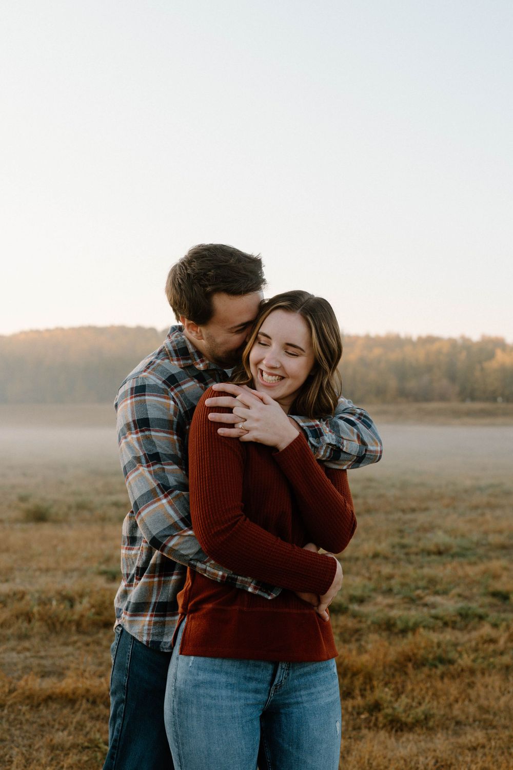 A couple embracing in a misty field at sunset wearing plaid and burgundy sweater.