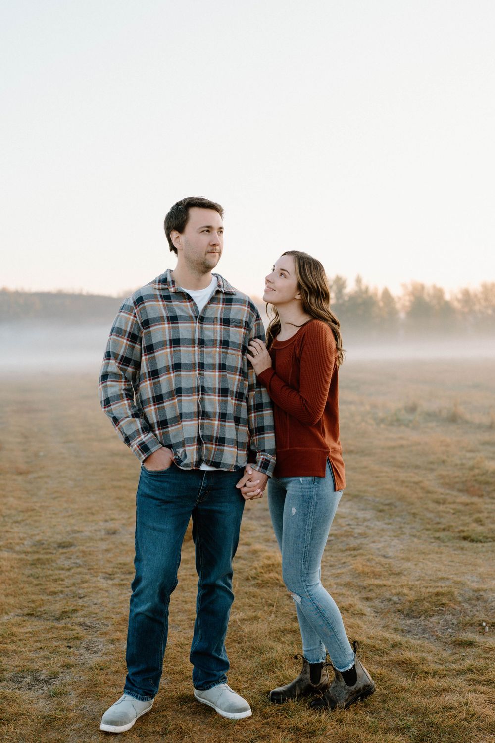 Couple in casual wear holding hands while standing in a misty morning field.