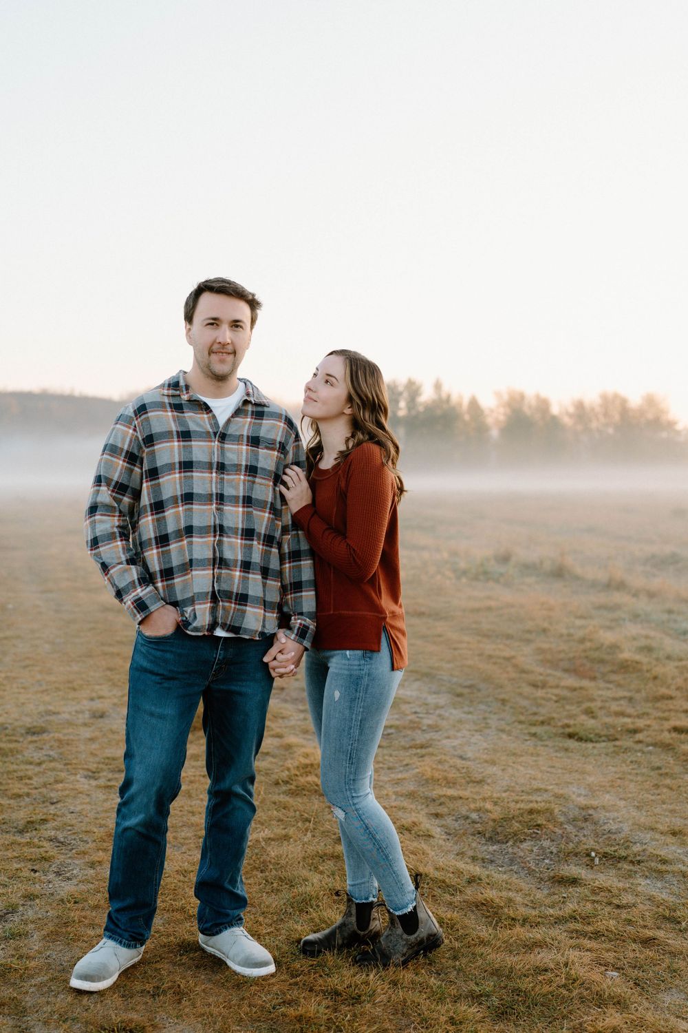 Couple in matching casual outfits standing together in a foggy field at sunrise.