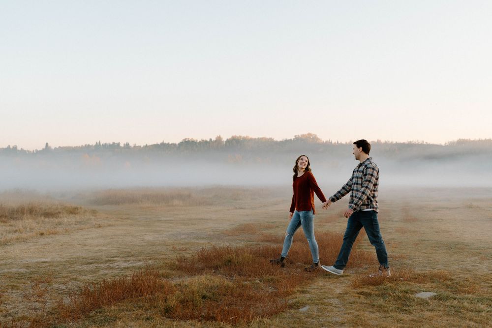 A couple holding hands while walking together in a foggy meadow at sunrise.
