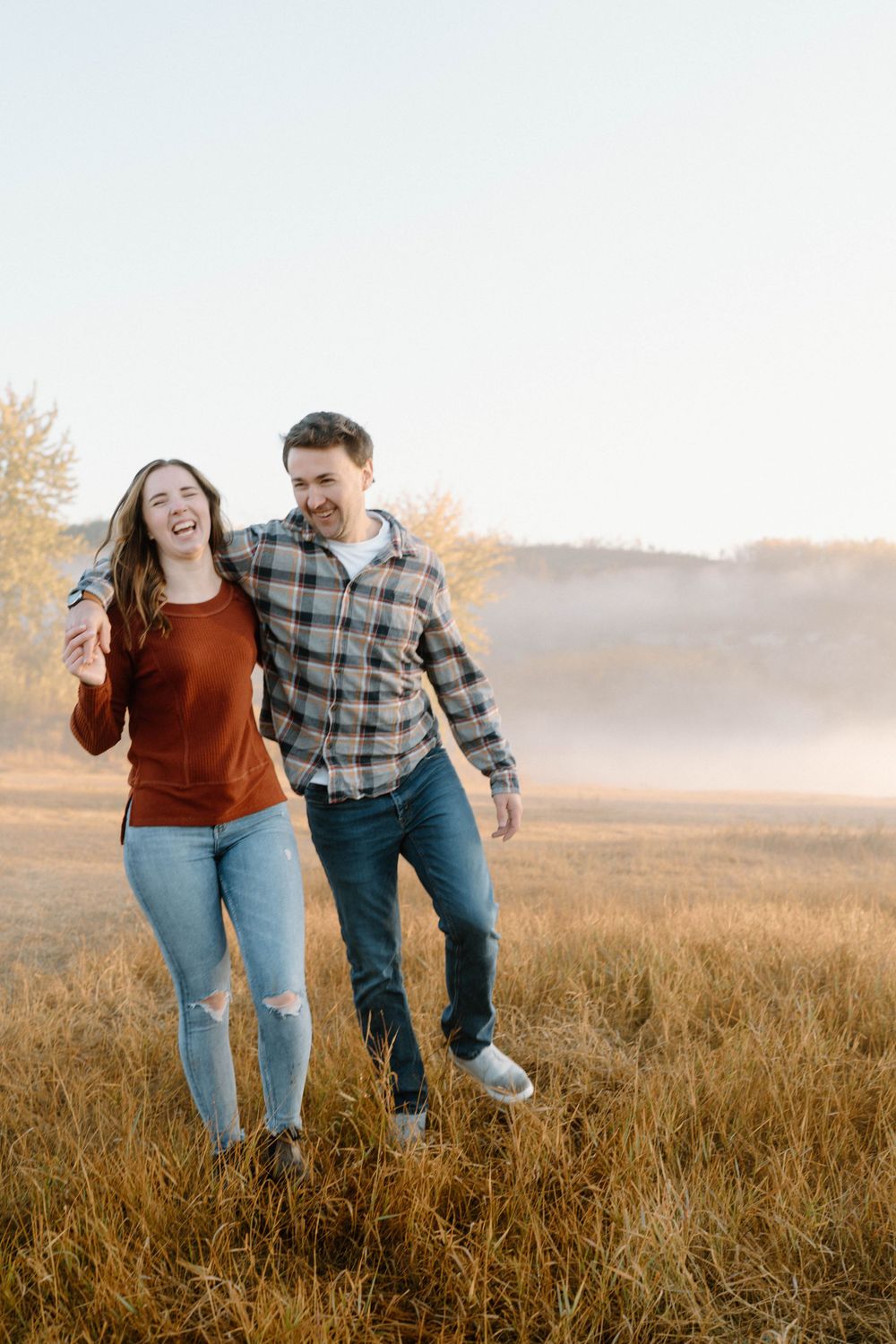 Couple laughing together while walking through a golden field at sunset in casual autumn attire.