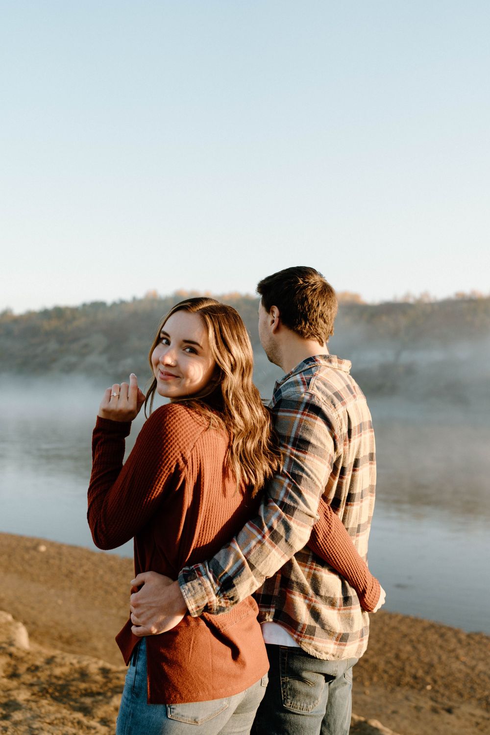 Two people enjoy a misty morning by a lake, wearing casual fall attire and sharing a playful moment.
