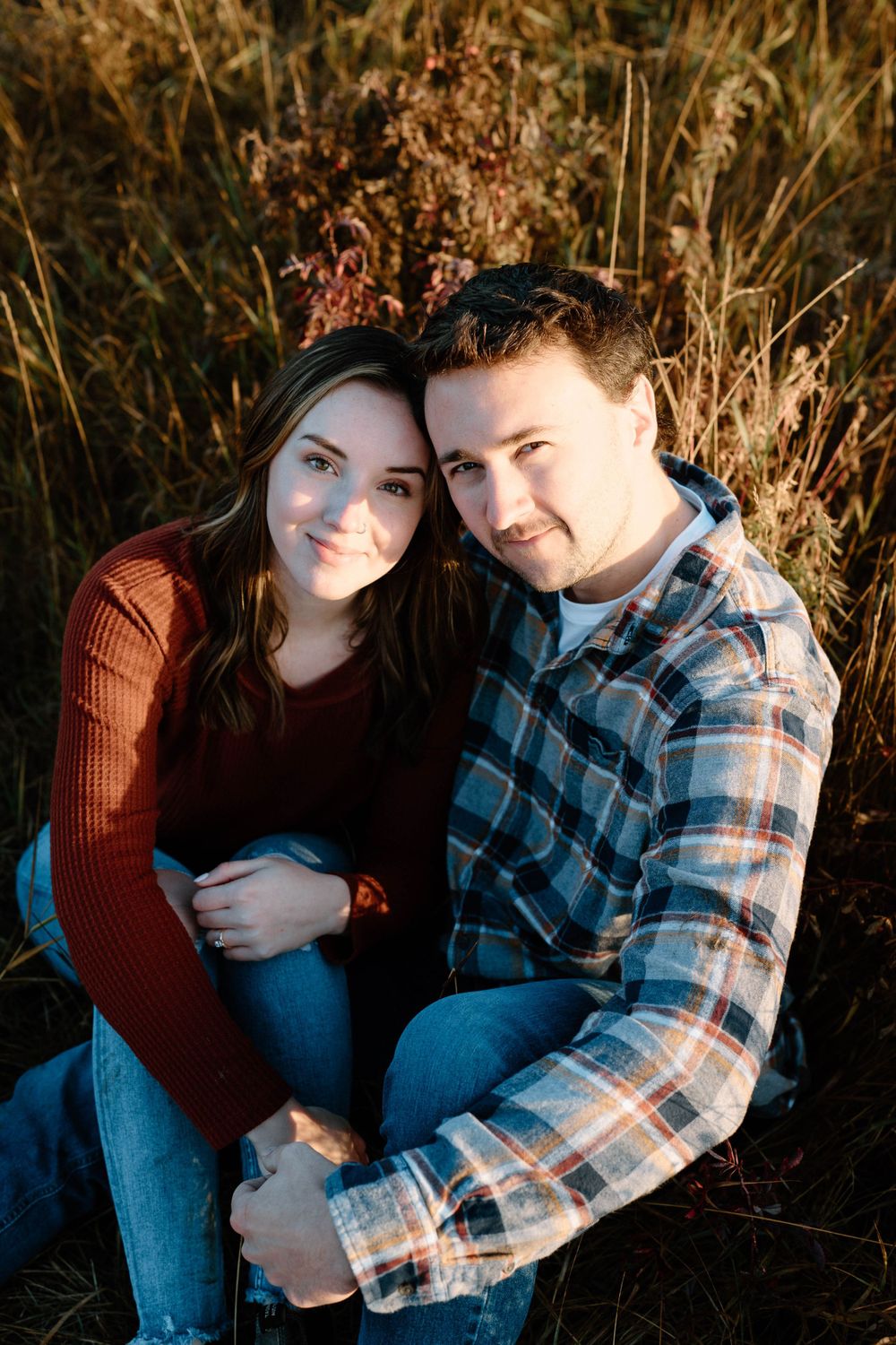 A couple in casual wear poses together amidst dried grass during golden hour.