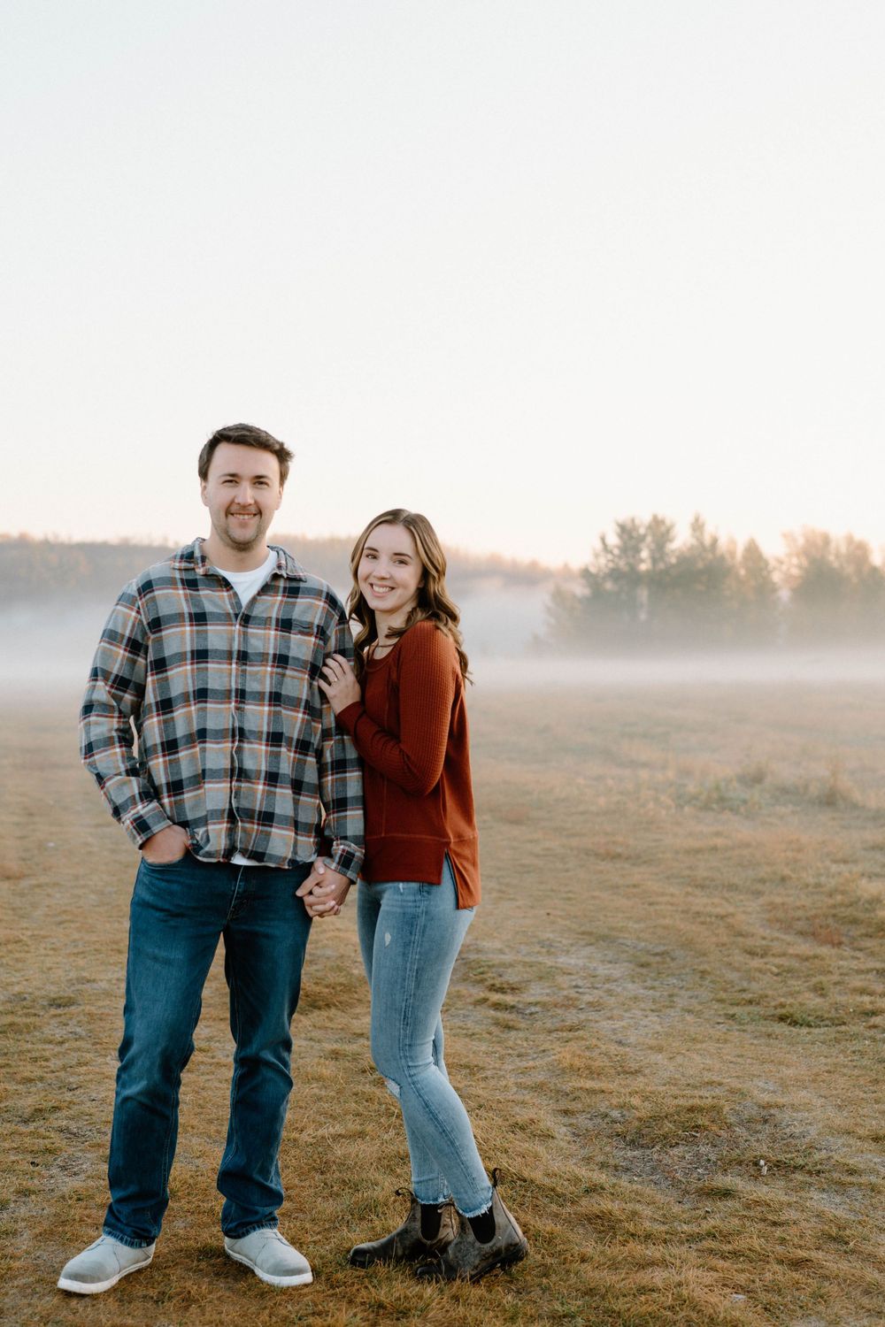Happy couple poses together in casual attire on a misty morning in a field.