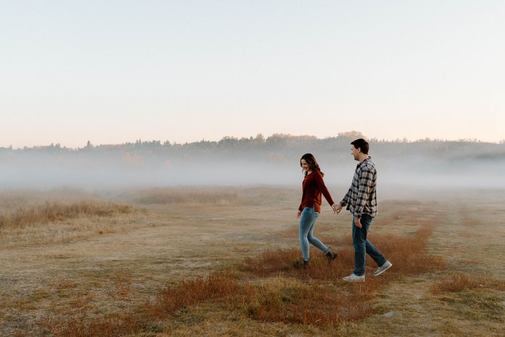 Couple walking hand in hand through a misty field during golden hour.