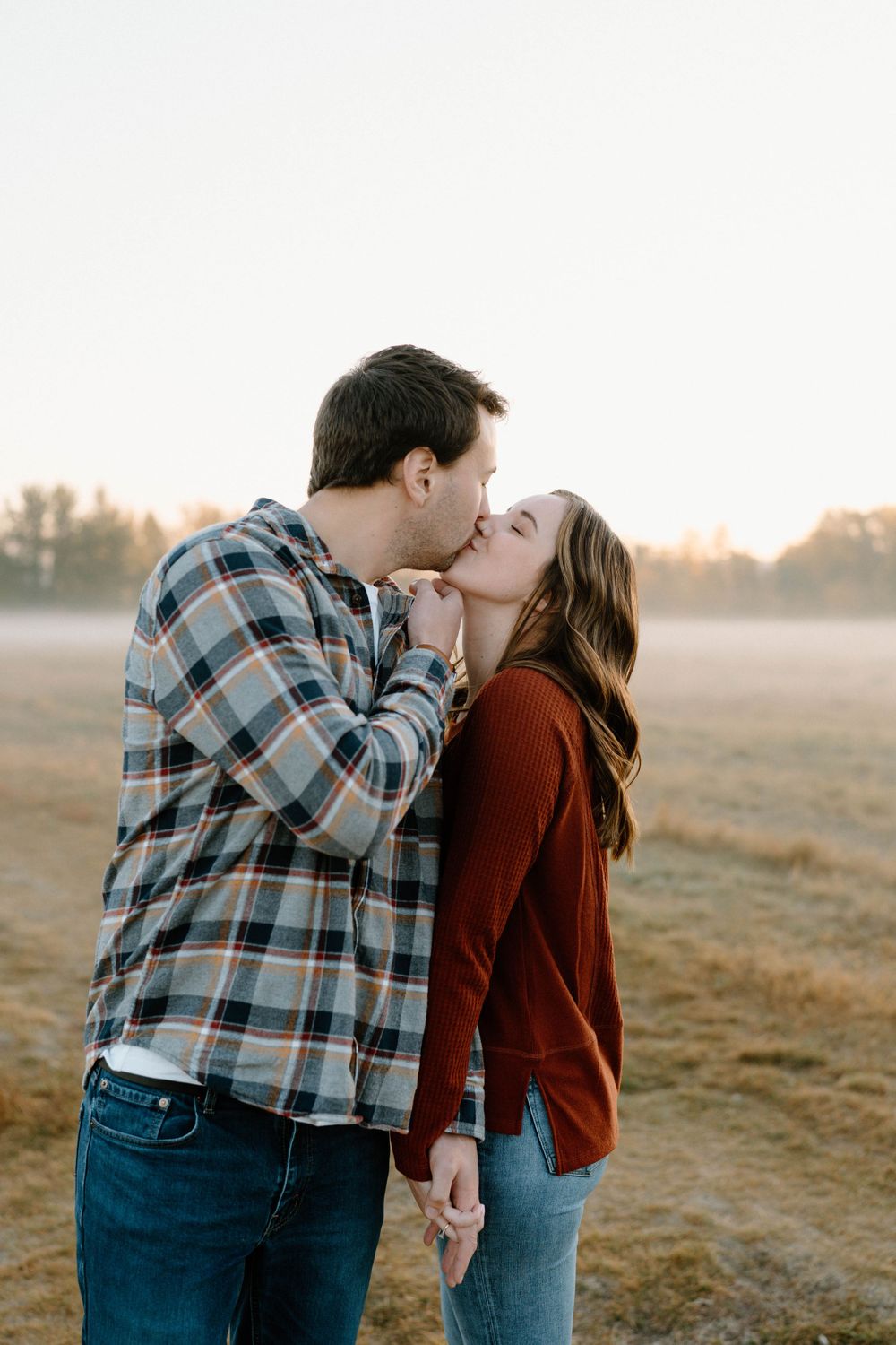 A couple shares an intimate kiss while standing in an open field at sunset.