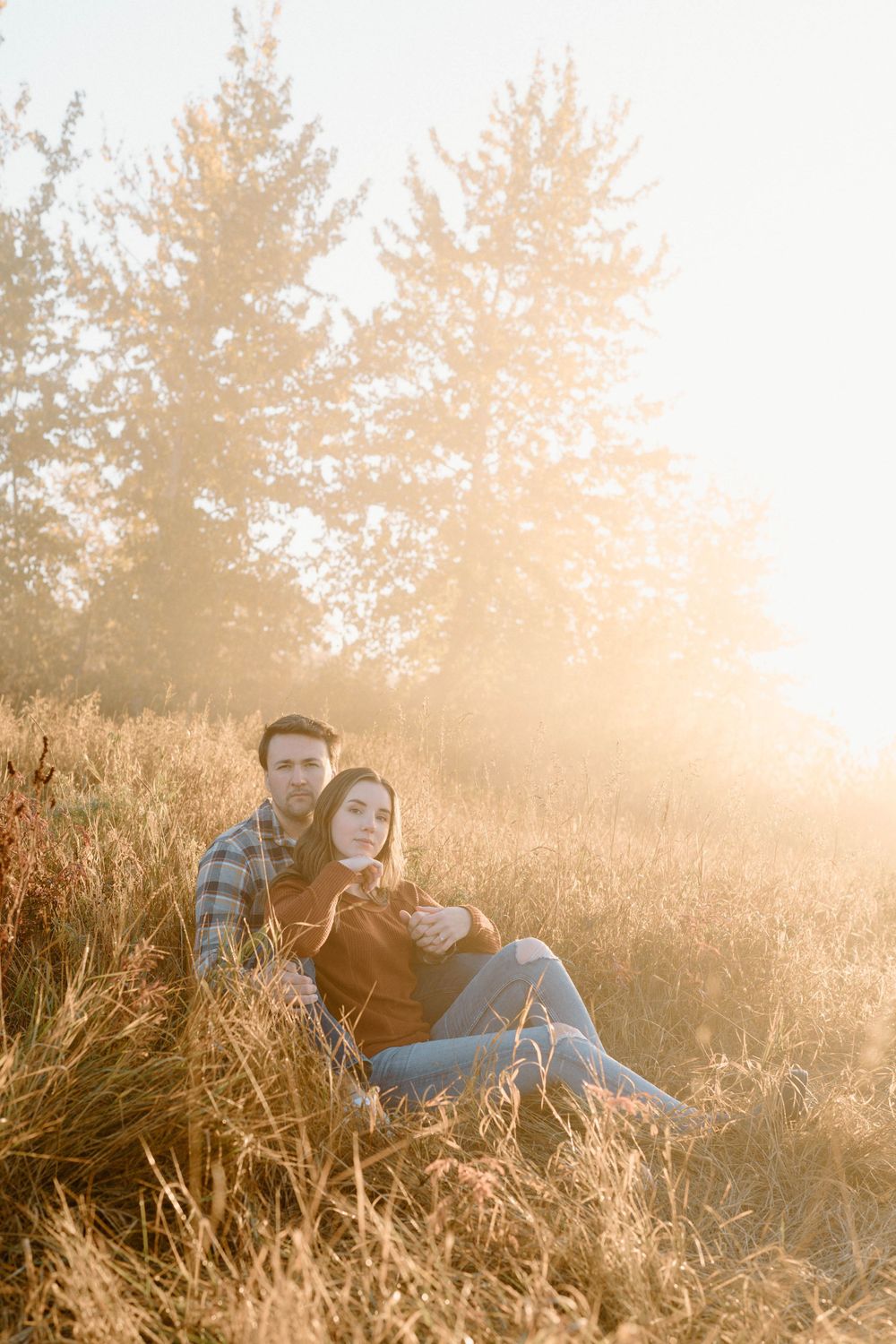 A couple sits together in golden sunlit grass with evergreen trees and warm sunlight creating a dreamy atmosphere.