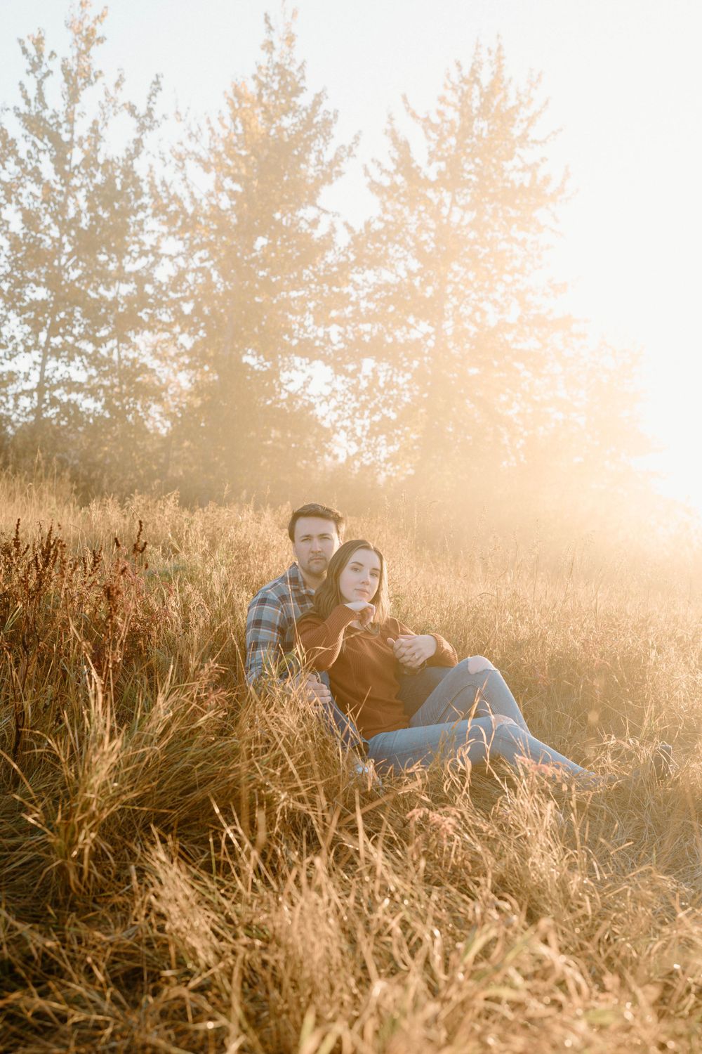 A couple relaxes in tall golden grass during sunset with pine trees in the background.