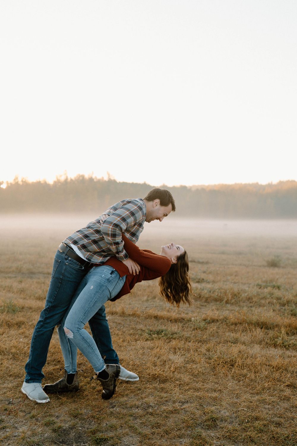 A playful couple in casual wear dips in a field during golden hour.