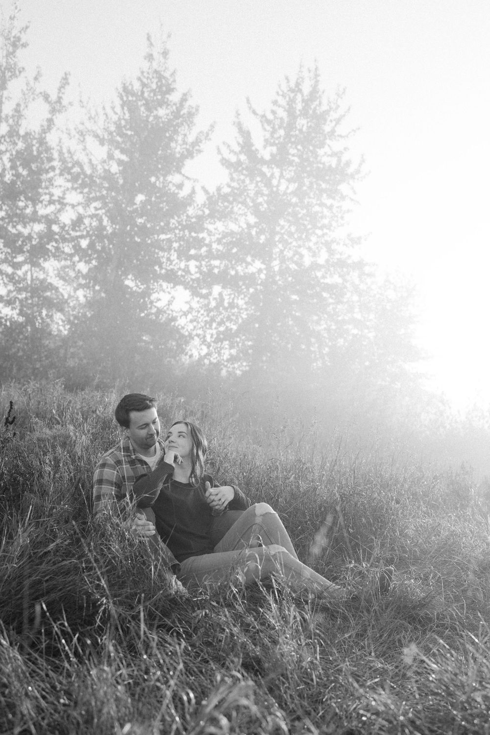 A black and white photo of a couple sitting together in a grassy field at sunset.