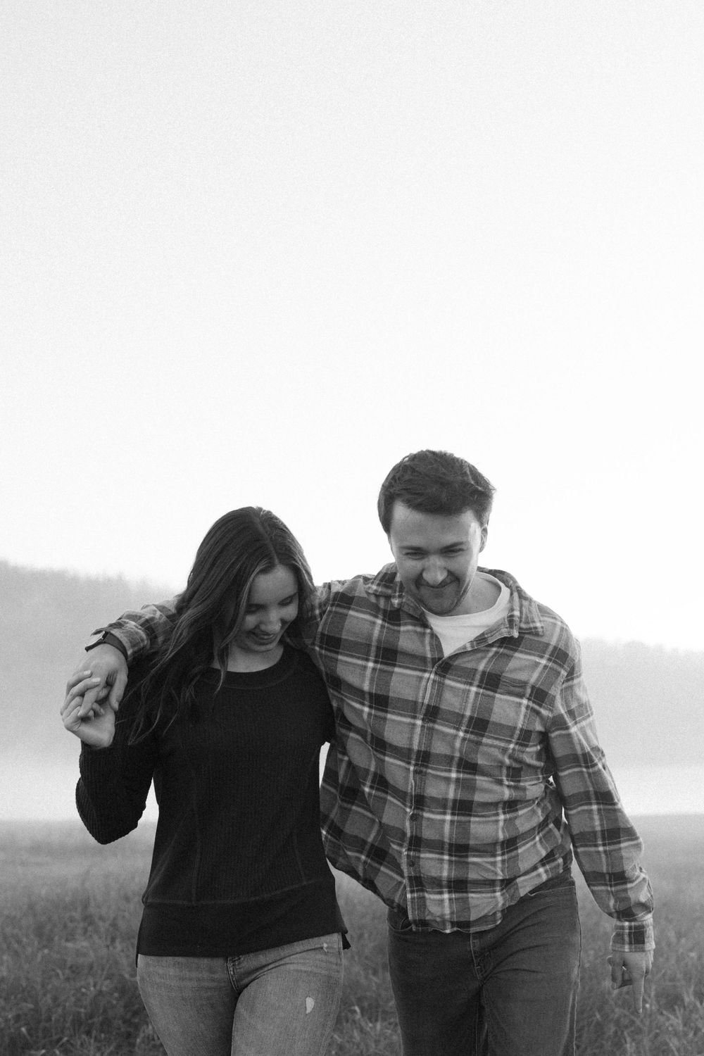 A black and white photo of a couple walking together and laughing in a misty field.