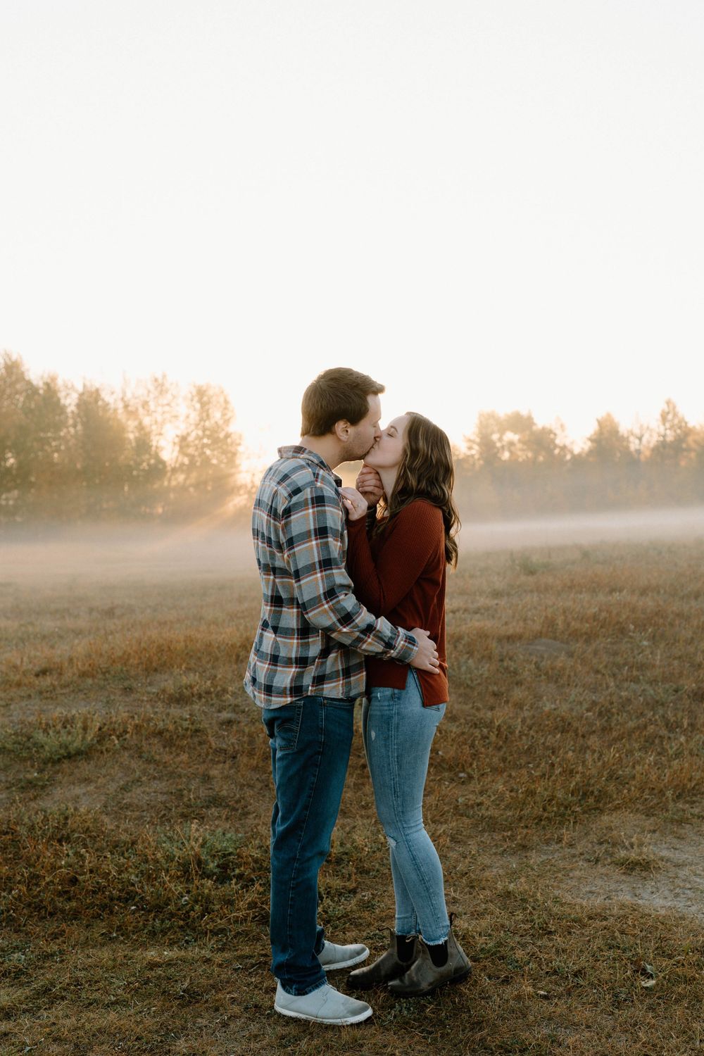 A couple in casual wear sharing an embrace on a foggy field at sunset.