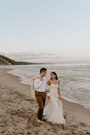 Couple walking along sandy beach at sunset in romantic wedding attire.