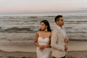 Couple in wedding attire poses back-to-back on beach during golden hour.