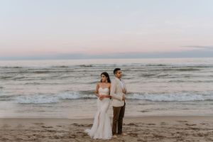 Couple in wedding attire standing on beach at dusk with waves rolling in.
