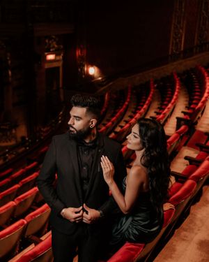 Glamorous couple in formal wear poses in historic theater with red velvet seats.