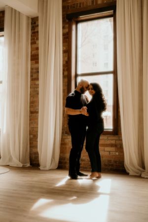 Couple dancing close together by a window in an elegant room with brick walls and white drapes.