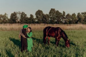 A couple in traditional attire poses in a meadow with a grazing horse at sunset.