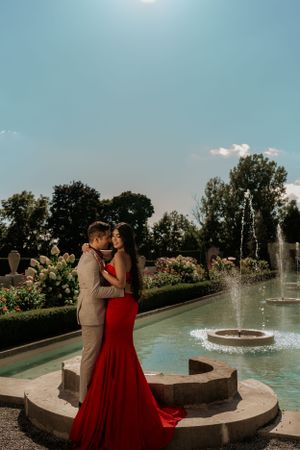 A romantic couple poses by a garden fountain with the woman wearing a flowing red gown and the man in a tan suit.