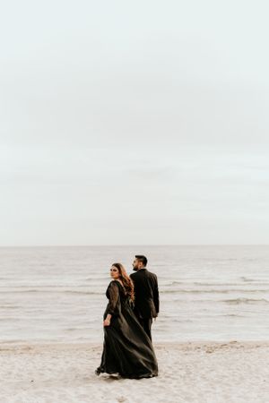Two people in dark formal wear stroll along a foggy beach shoreline.