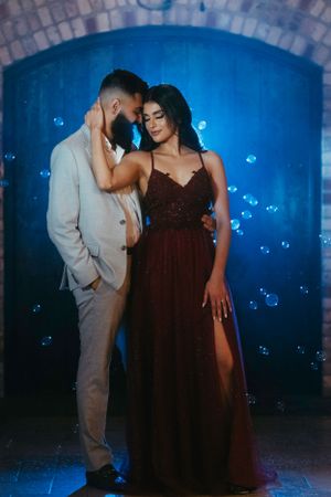 Couple in formal attire poses against blue-lit archway with floating bubbles.