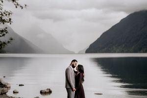 A couple poses intimately by a serene mountain lake with misty peaks in the background.