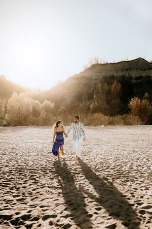 Couple walking hand in hand on sandy beach at sunset casting long shadows.