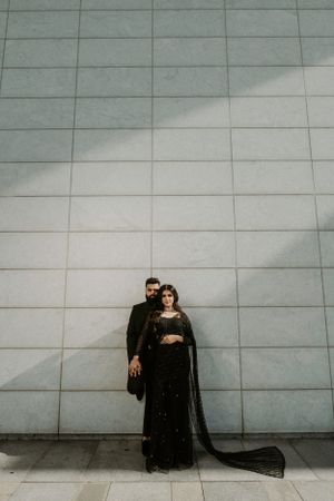 Couple in dark formal wear stands against minimalist tiled wall with striking shadows.