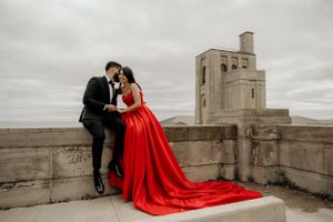 A dramatic wedding portrait featuring a long red dress against concrete architecture under cloudy skies.
