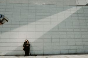 Figure in black standing alone against large white tiled wall with diagonal sunlight.
