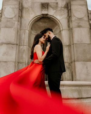 A romantic couple embraces under a stone archway with a flowing red dress creating a dramatic effect.
