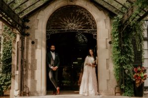 Two people stand apart in an ornate archway covered with lush green vines.