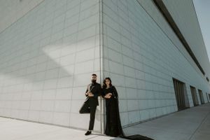 Elegant couple in black attire pose against a modern white geometric building wall.