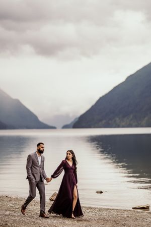A couple walks along a misty lakeside with mountains creating a dramatic backdrop.