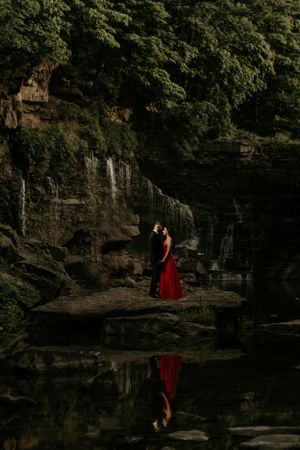 Couple in formal attire embracing near a waterfall with stunning reflection in the still water.