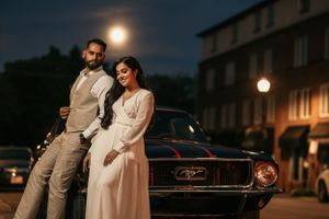 A stylish couple poses against a classic car on a moonlit street with vintage architecture.