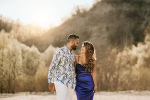 Stylish couple in blue dress and patterned shirt pose together at sunset in a natural setting.