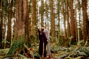 Two figures embrace in a majestic Pacific Northwest forest surrounded by towering evergreen trees and lush ferns.