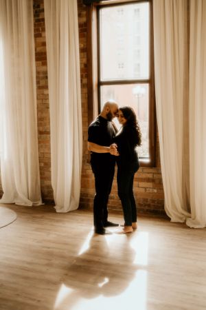 A romantic couple shares an intimate dance in front of a sunlit window with flowing white curtains and exposed brick.
