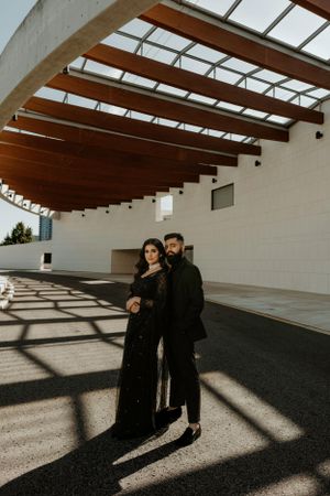 Couple in dark attire under geometric wooden ceiling with dramatic shadow patterns.