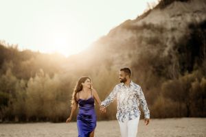 Couple in blue dress and white patterned shirt walking together at golden hour in mountains.