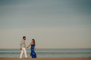 Couple in formal attire holding hands while walking along the beach at sunset.