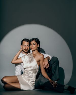 Stylish couple in modern formal wear poses intimately against dramatic circular spotlight backdrop.