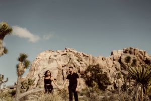 Couple posing against dramatic desert rock formations and Joshua trees at sunset.
