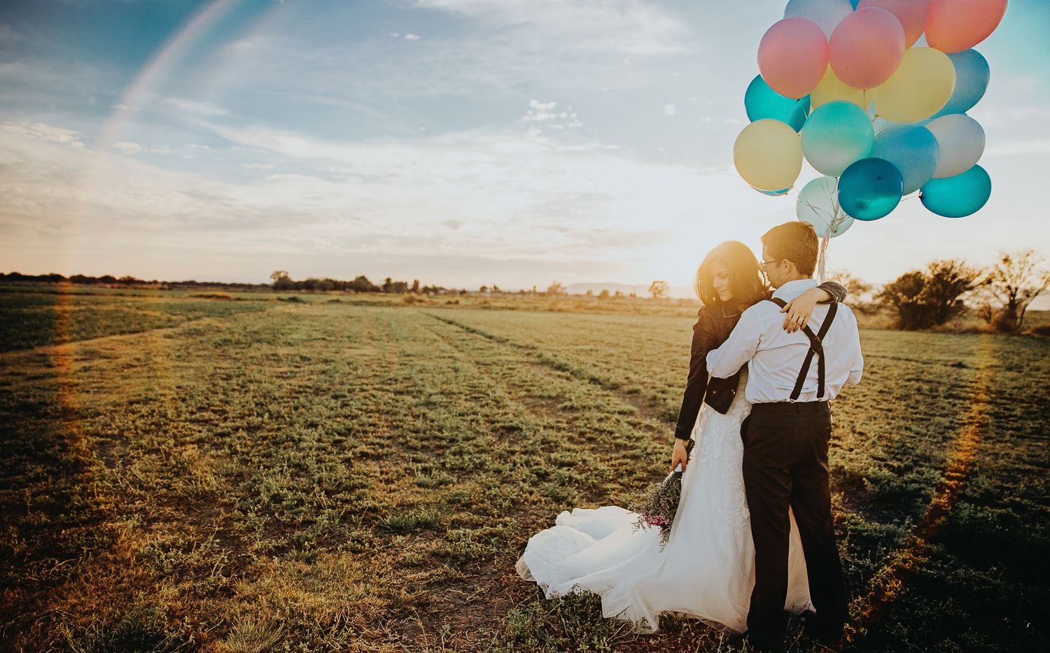 A wedding couple holds colorful balloons in a field at sunset.