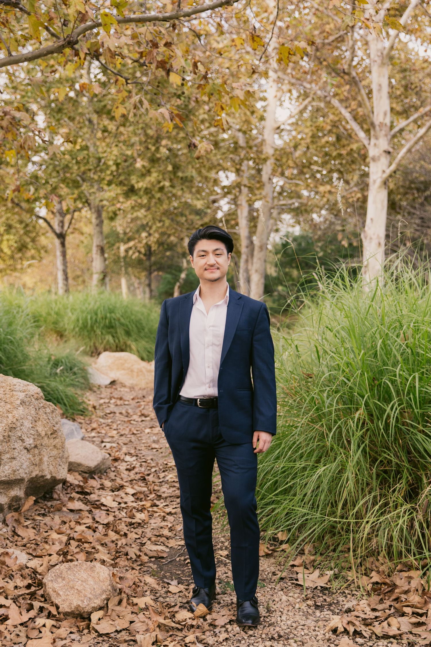 A person in a navy suit poses casually on a rocky path surrounded by autumn trees.