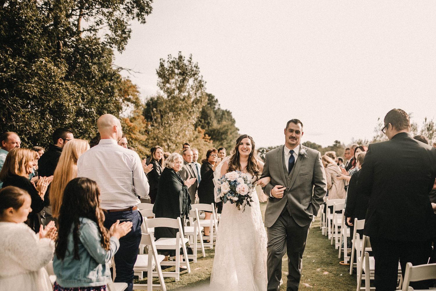 Wedding guests gather outdoors with white chairs at a charming ceremony venue in warm lighting.
