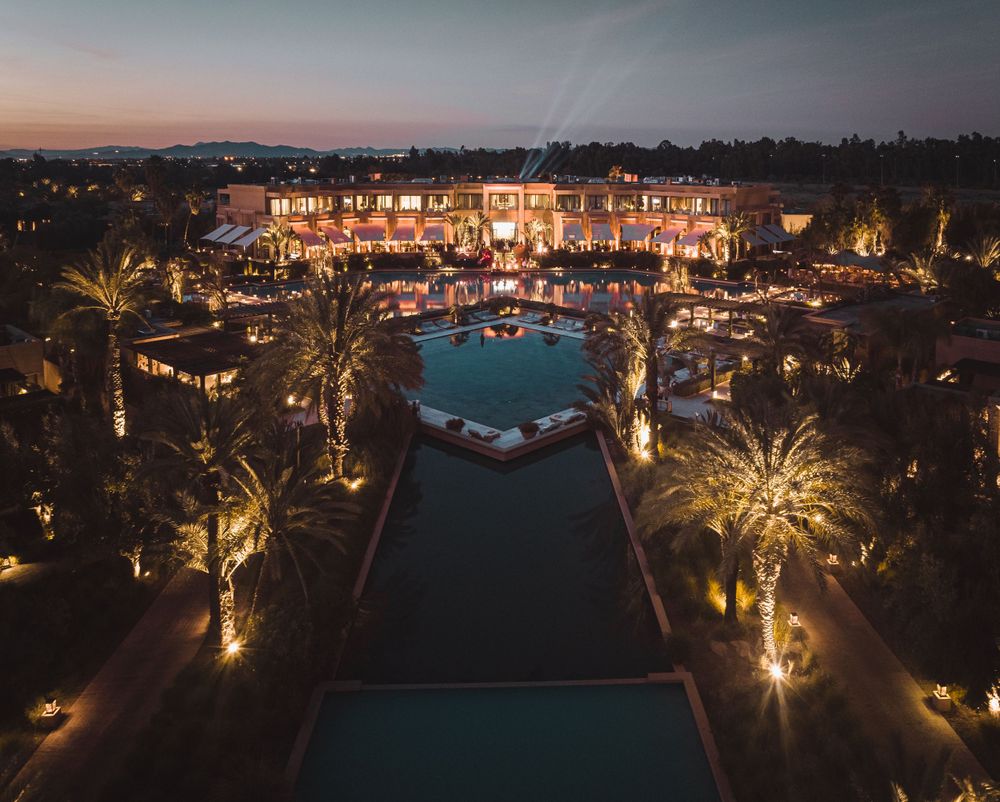 Aerial view of a luxurious resort with illuminated palm trees and a central swimming pool at dusk.